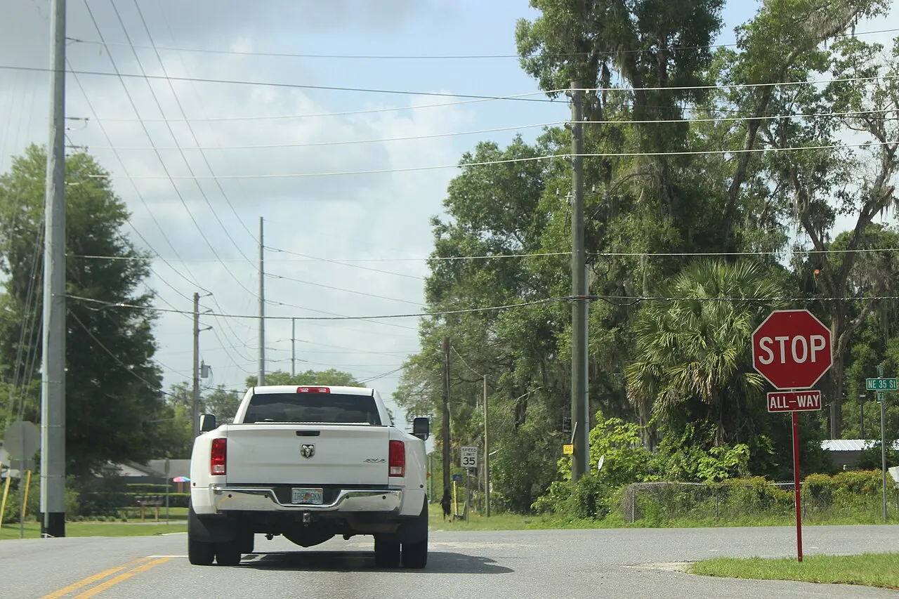 Stop sign with a red ALL-WAY plaque on the right side of a two-lane road as a pickup truck approaches
