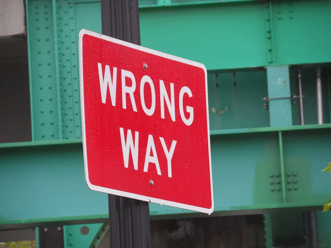 Red Wrong Way sign with white lettering on a pole in front of green steel girders in Boston