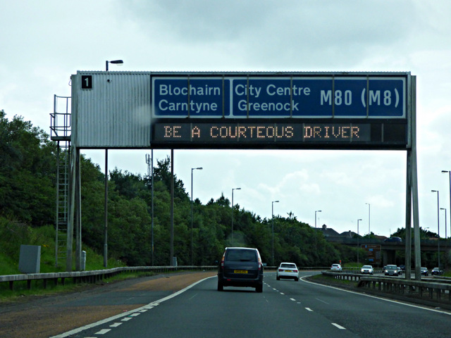 A motorway gantry sign on the M80, evoking the dense junction-heavy traffic that defines London test centre routes